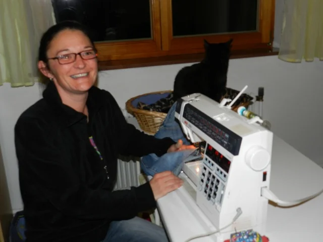 Eine Frau sitzt lächelnd an einer Nähmaschine und bearbeitet ein Kleidungsstück.